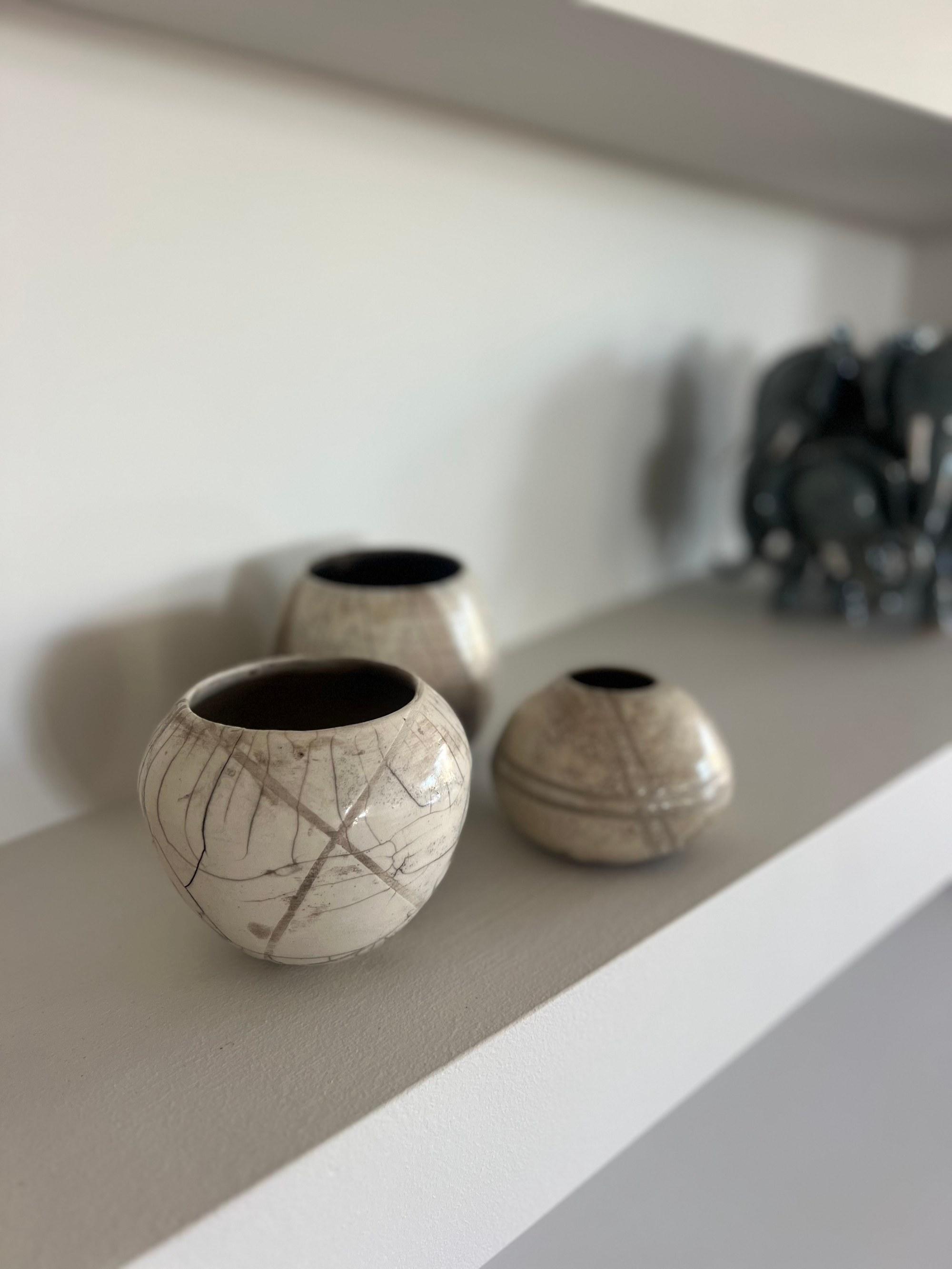 Three pale raku-fired pots on a shelf, crazed white with smoke-grey lines, an elephant herd behind them