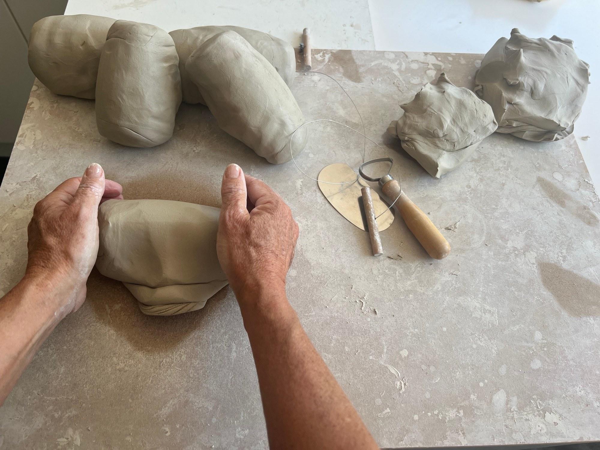 Louise's hands shaping a block of clay on a stone worktop, with a wire cutter and modelling tools beside her